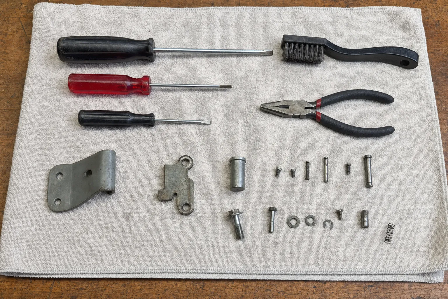 Screwdrivers, pliers, a wire brush and a hinge bracket with its fasteners laid out on a shop towel